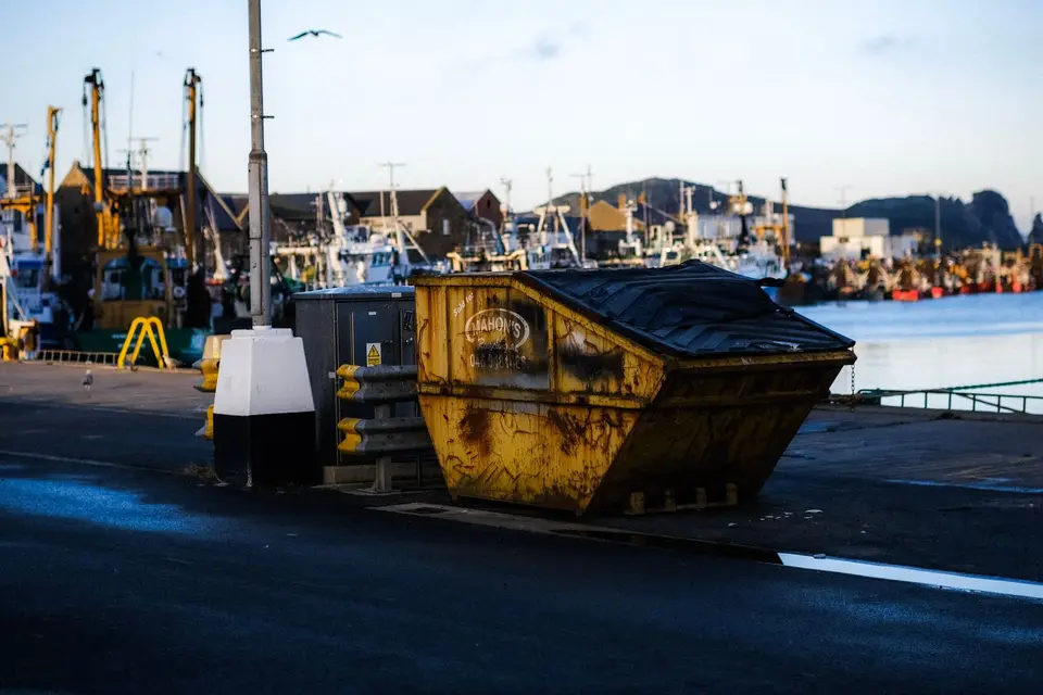 Benne jaune posée sur un quai, devant des bateaux de pêche